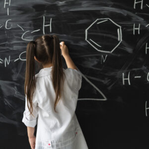 Cute little girl writing on a blackboard at a chemistry lesson in class. White coat and goggles.